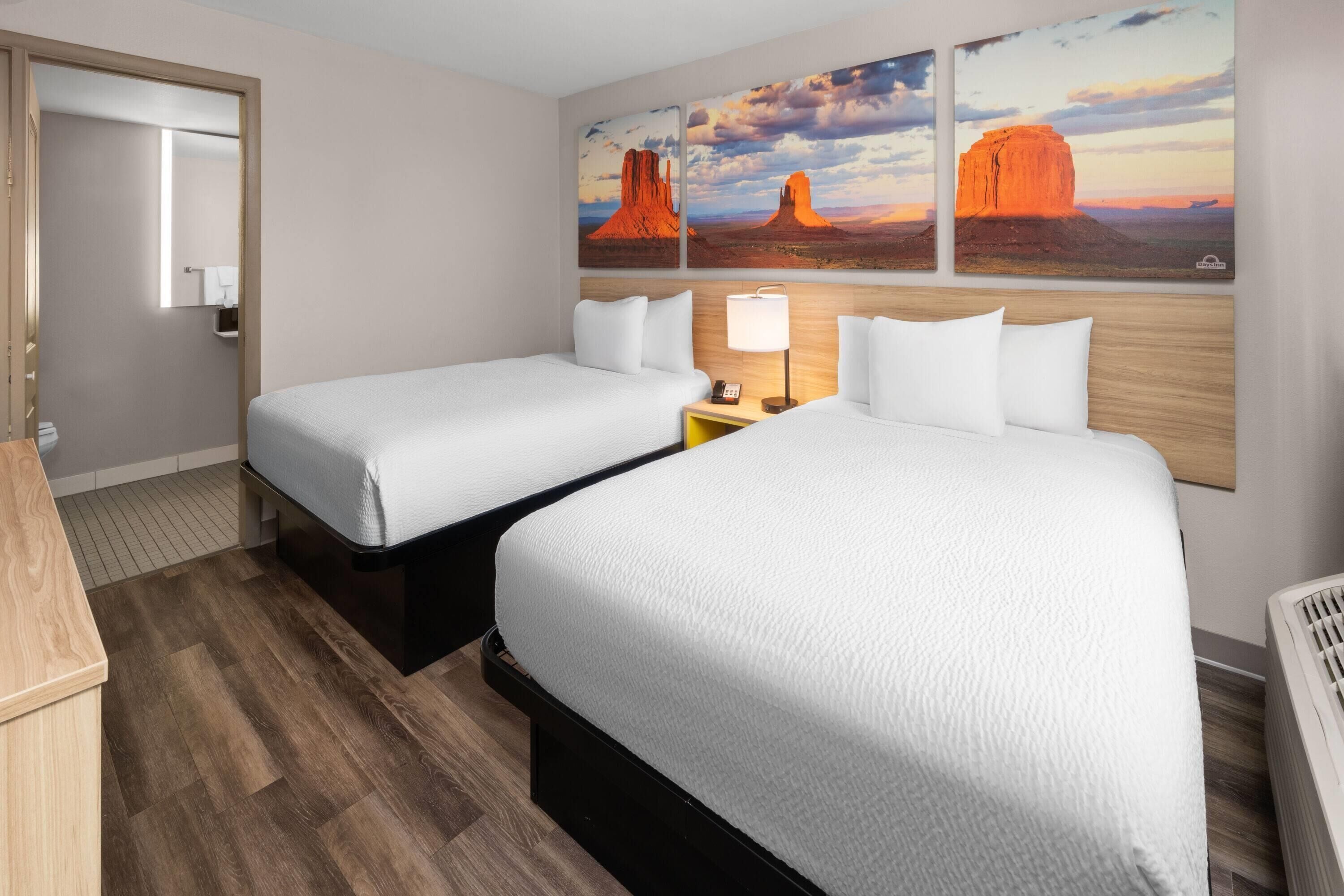 Habitación de hotel con dos camas, piso de madera, mesita de noche con lámpara y decoración mural con temática desértica.