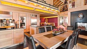 Une cuisine moderne dotée d’appareils électroménagers en acier inoxydable, un coin repas avec une table en bois et des chaises en cuir noir, et une cheminée surmontée d’une horloge.