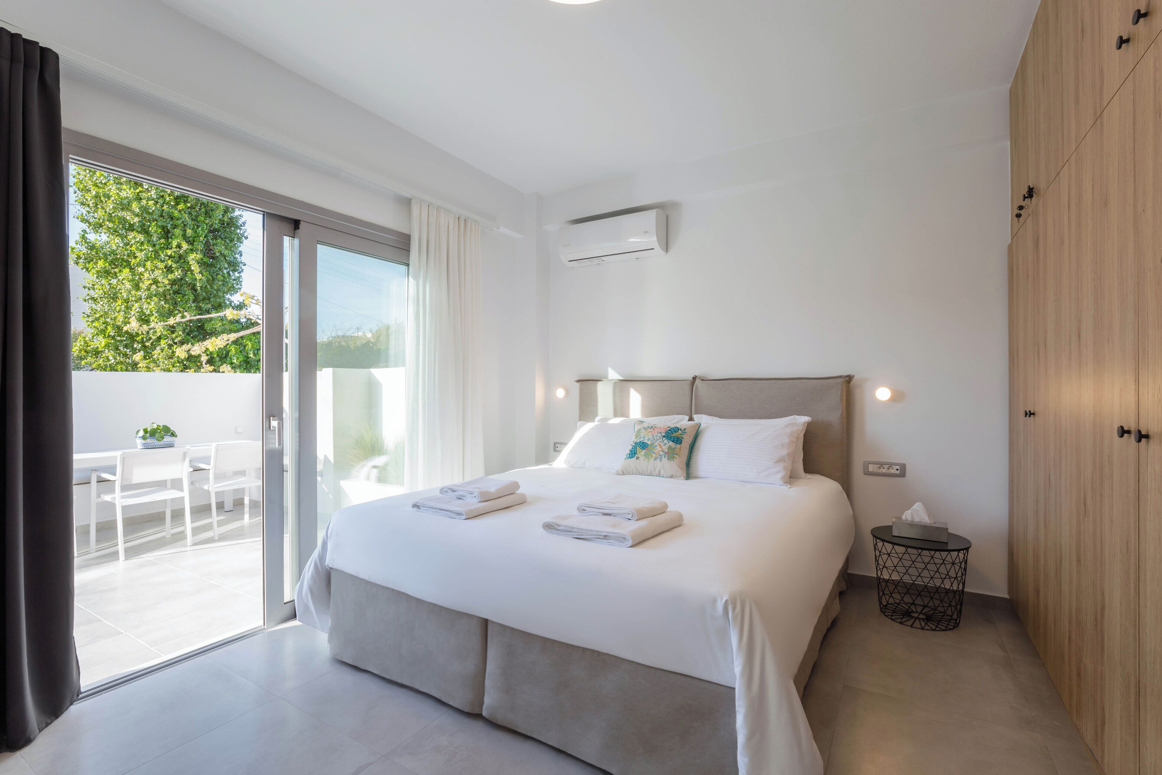 A modern bedroom with a large bed, a sliding glass door to a balcony, and a wooden wardrobe.