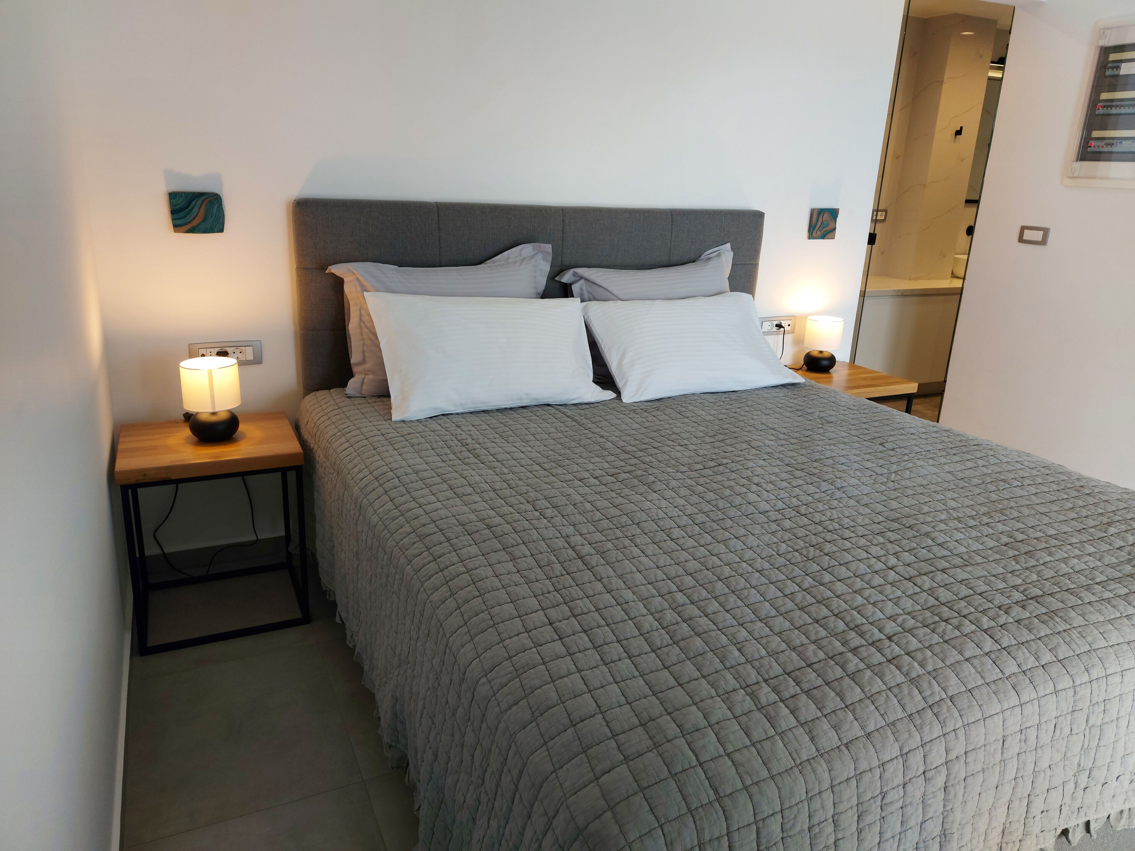 A neatly made bed with a grey quilt, two white pillows, and a grey headboard. There are two bedside lamps on wooden nightstands.