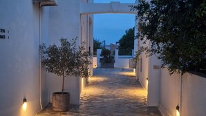 Ένα στενό σοκάκι με λιθόστρωτο δρόμο, λευκούς τοίχους, ένα φυτεμένο δέντρο και ένα κτίριο με μια εξωτερική μονάδα κλιματισμού.