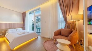 Quarto de hotel moderno com uma cama grande, um espaço de estar com uma poltrona, televisão e varanda com cortinas.