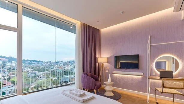 Quarto de hotel com uma cama grande, vista para a cidade, parede roxa, televisão de tela plana, luminária e uma cadeira.