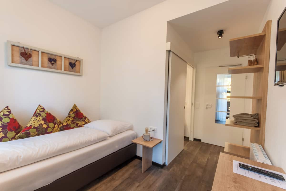 A bedroom with a bed, pillows, a bedside table, a wall-mounted shelf, and a door leading to another room.