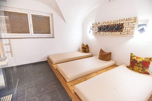 A room with three beds, a wooden bench, a window with blinds, and a wall-mounted decorative piece. - Pension Max (Zell am See)