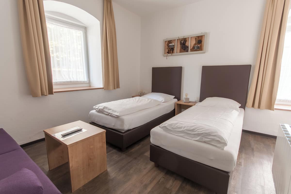 A hotel room with two single beds, a wooden nightstand, a small table, a purple sofa, and a window with curtains.