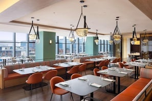 A modern restaurant with high tables and orange chairs, large windows offering city views, and unique hanging light fixtures.