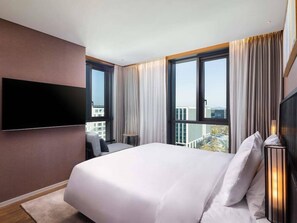 Ein Hotelzimmer mit einem großen Bett, einem an der Wand befestigten Flachbildfernseher, einem Fenster mit Vorhängen und Blick auf die Stadt.
