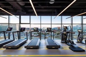 Een sportschool met loopbanden, elliptische trainers en een roeimachine, grote ramen en uitzicht op de stad.