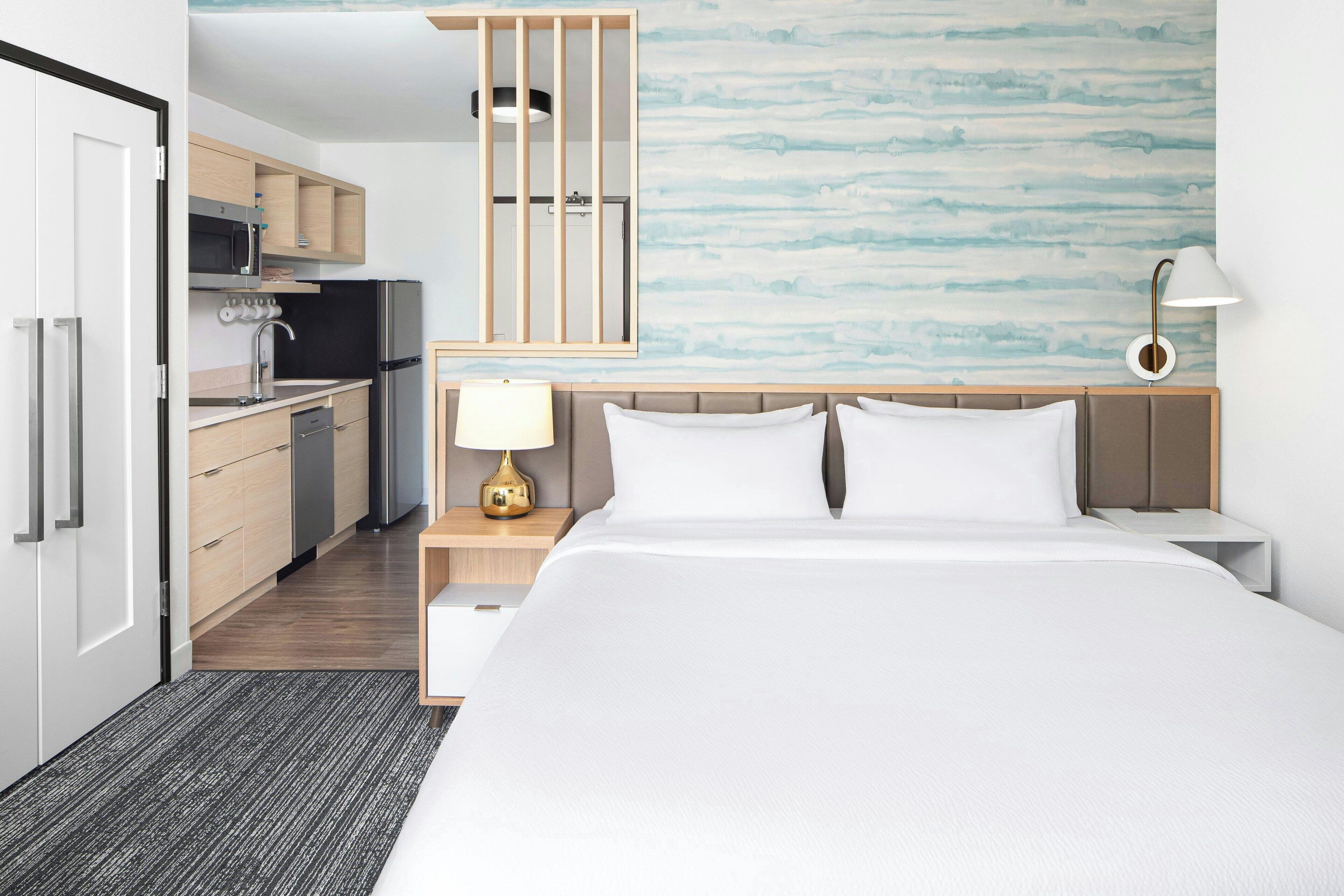 Ein modernes Hotelzimmer mit Bett, Nachttisch, Lampe und Blick zur Küche.