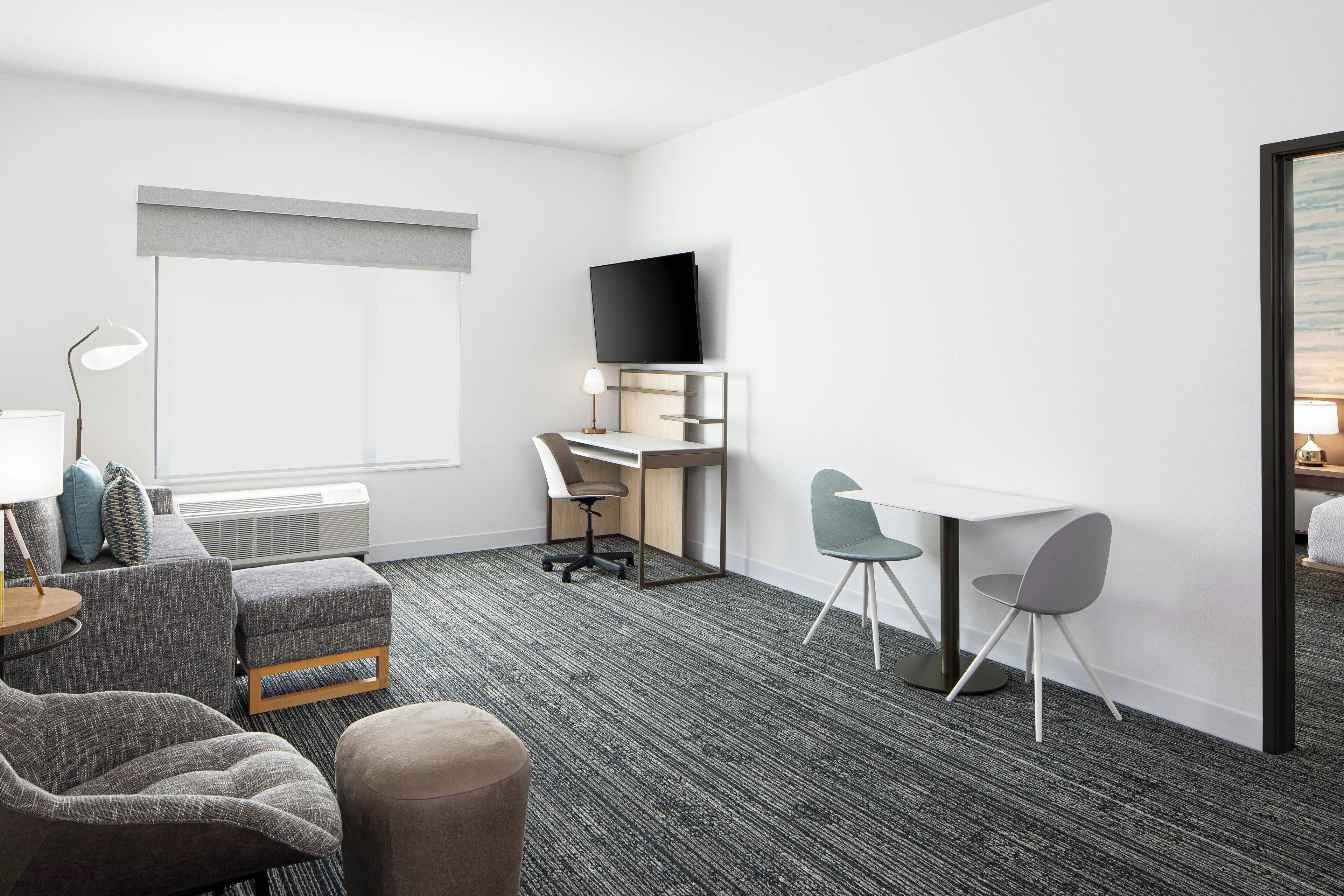 A hotel room with a grey carpet, a sofa, a desk with a chair, a TV mounted on the wall, and a window with a view.