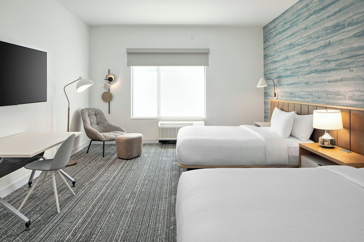Quarto de hotel com uma televisão de tela plana grande, uma escrivaninha com cadeira, duas camas com cabeceiras, ar-condicionado fixado na parede e uma janela grande com vista.