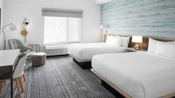 Quarto de hotel com duas camas, uma escrivaninha, uma cadeira, um abajur e uma janela grande.