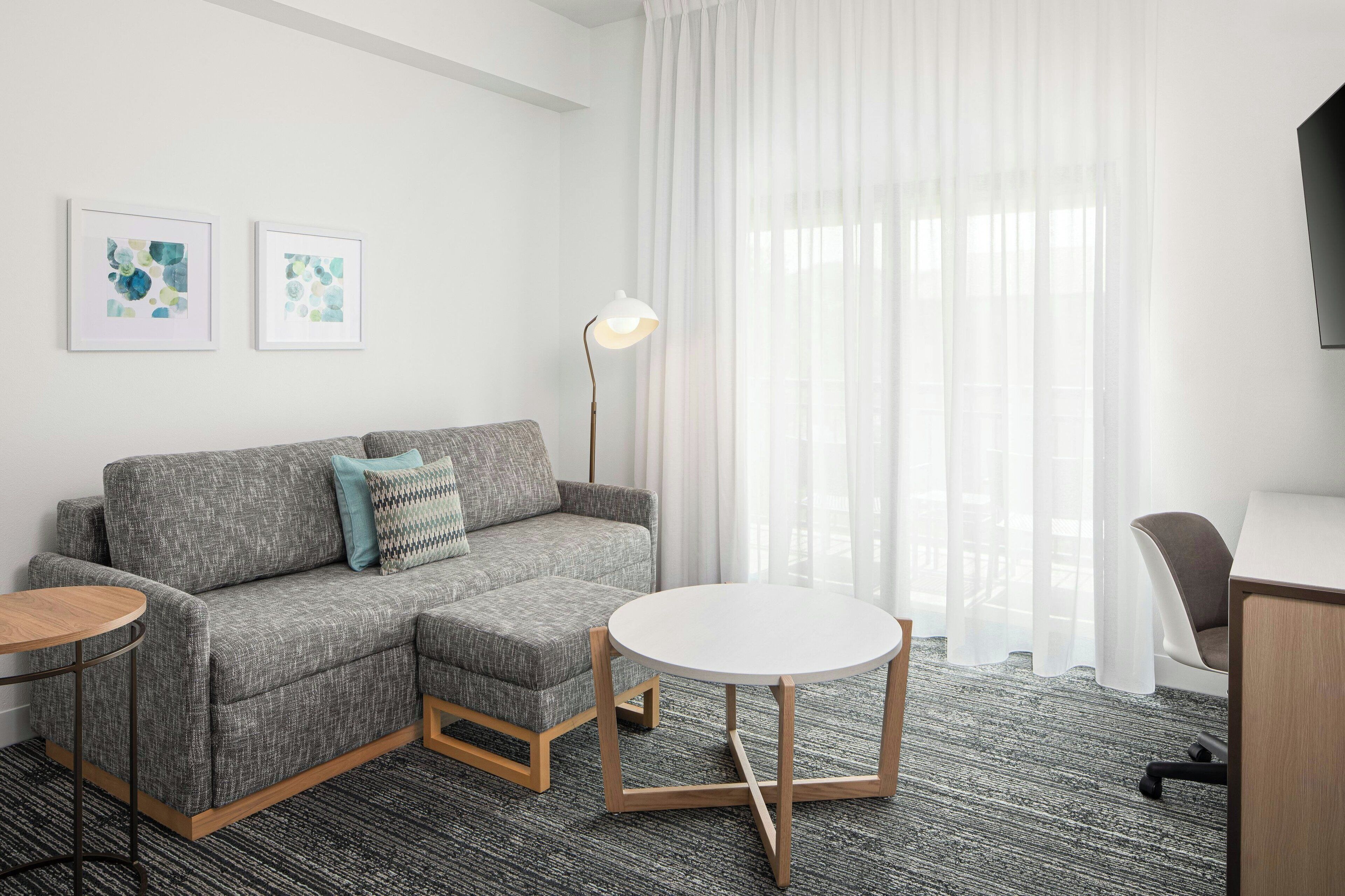 Une chambre d’hôtel moderne avec un canapé gris, une table ronde, un bureau et une grande fenêtre ornée de rideaux légers.