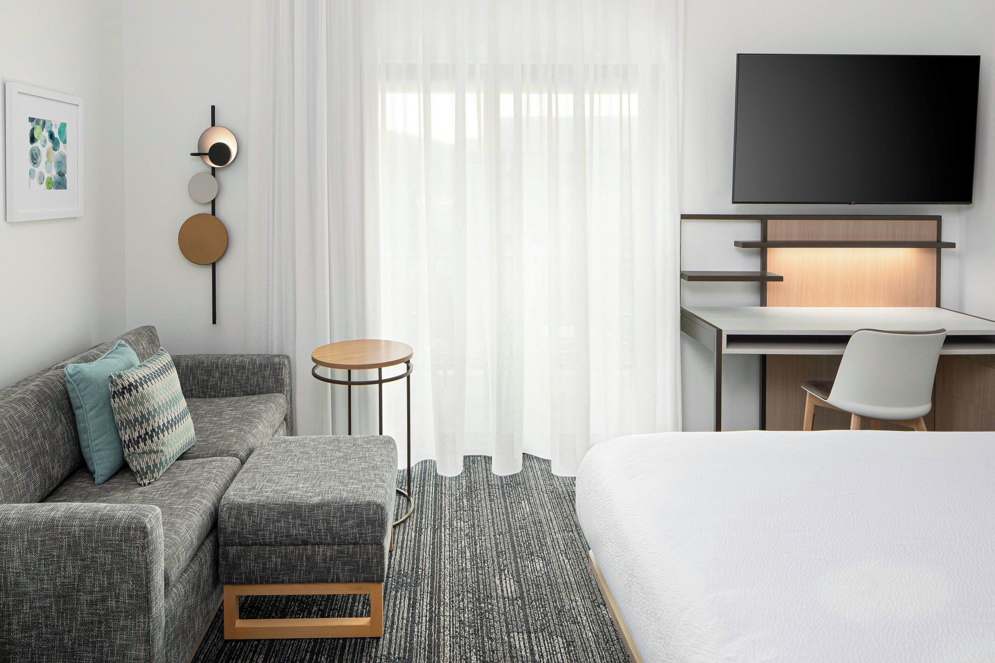 Une chambre d’hôtel moderne avec un canapé gris, une petite table ronde, un lit, un bureau avec une chaise, un téléviseur fixé au-dessus du bureau et un tableau encadré au mur.