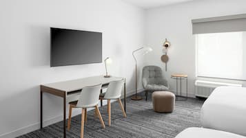 A modern hotel room with a flat-screen TV, a desk with two chairs, a grey armchair, a small round table, and a large window with a view.