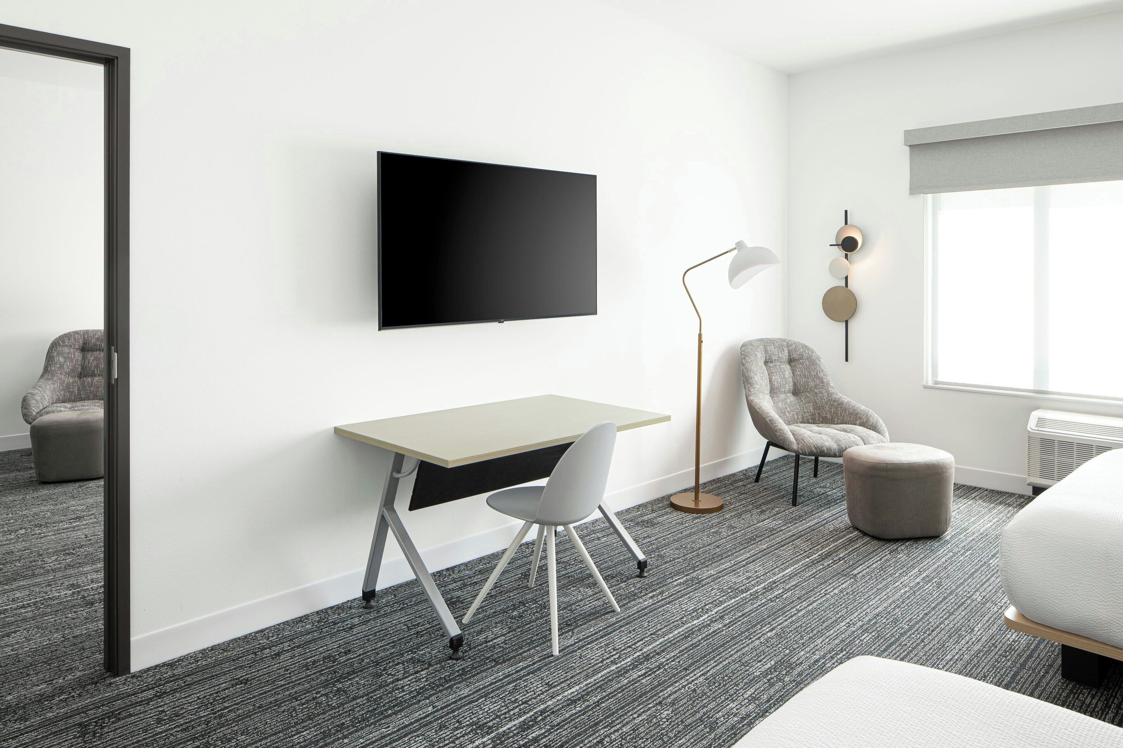 Une chambre d’hôtel moderne équipée d’un téléviseur à écran plat fixé au mur, d’un bureau avec une chaise, d’une lampe sur pied, d’un fauteuil gris, d’un pouf rond et d’un grand lit.