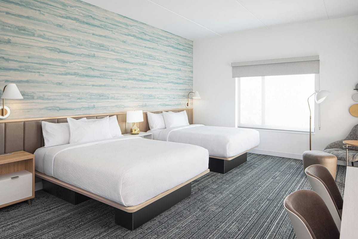Quarto de hotel com duas camas, uma janela grande, parede com design texturizado e um pequeno espaço de estar.