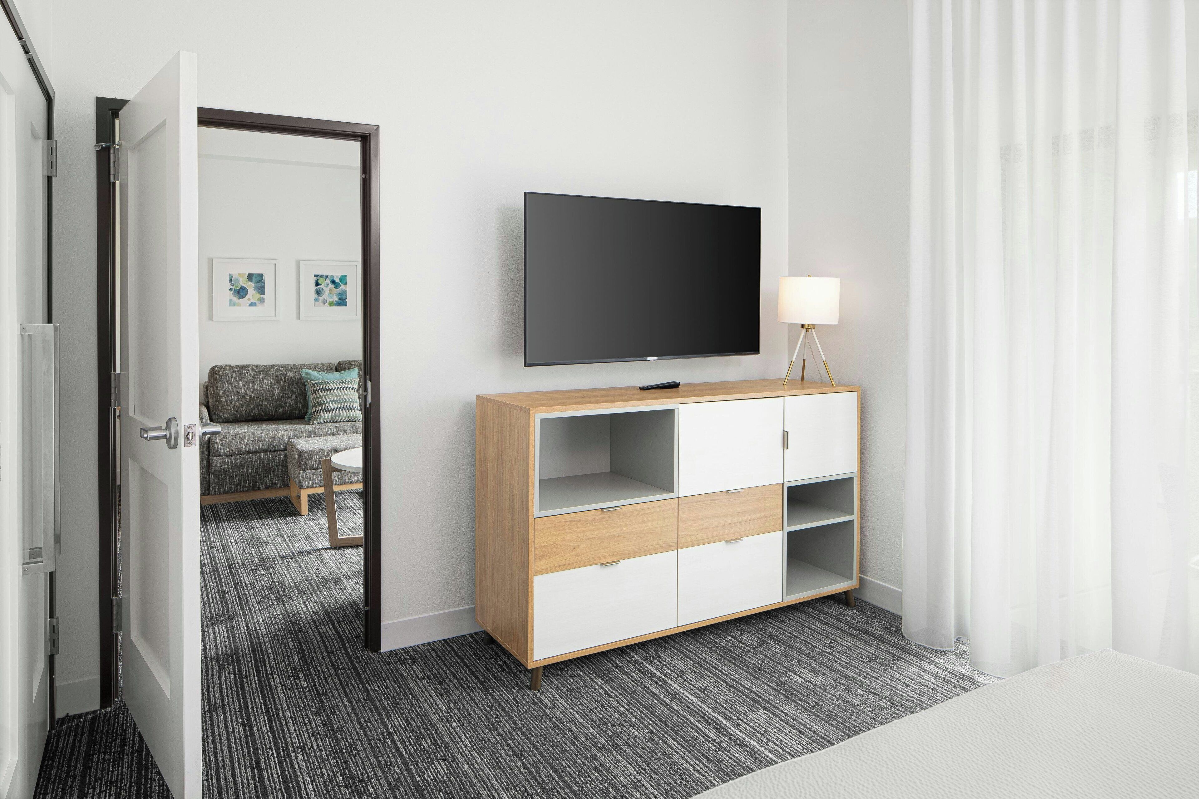 A hotel room with a large flat-screen TV mounted on the wall, a wooden and white cabinet with drawers, a small round table with a lamp, and a grey carpeted floor.