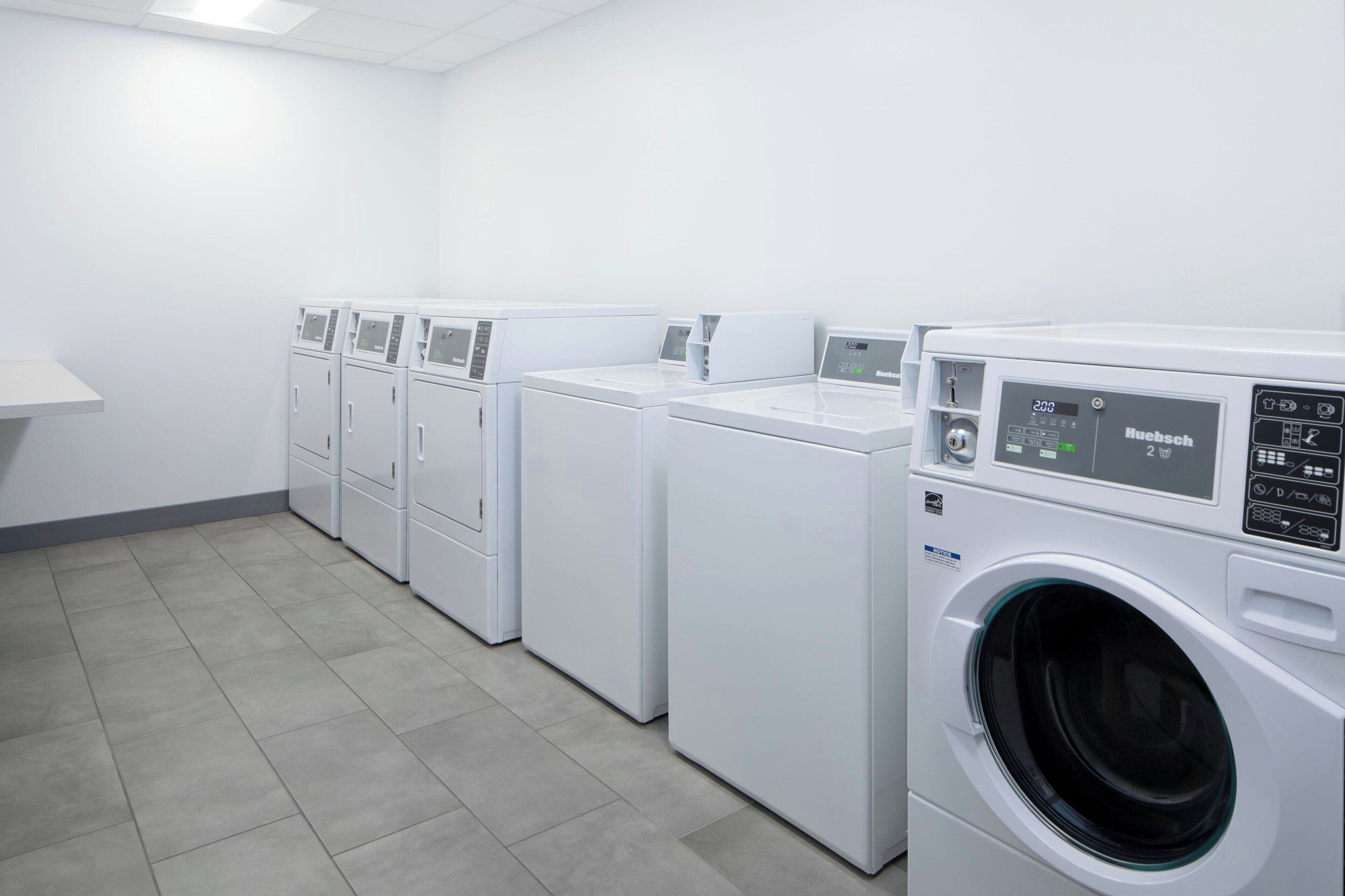 Une buanderie avec plusieurs machines à laver et sèche-linge blancs, un sol carrelé et un mur blanc.