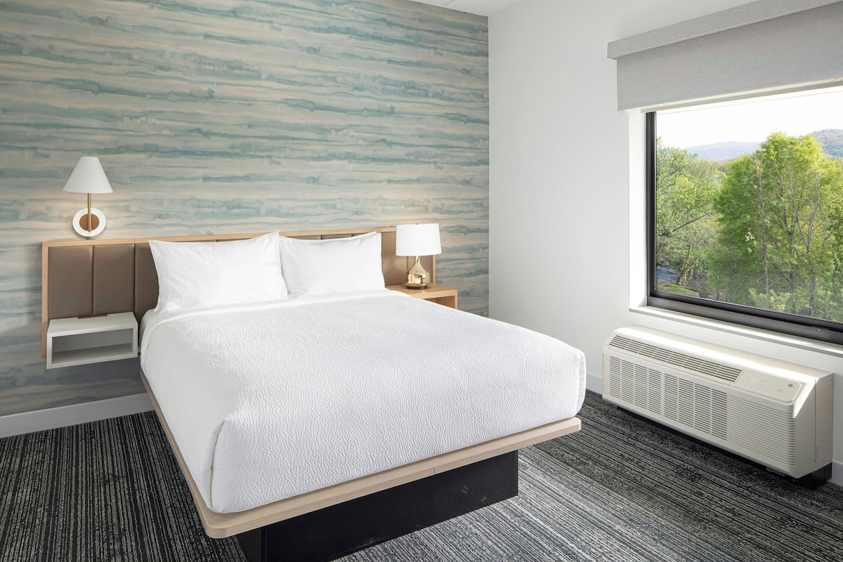 Habitación de hotel con una cama grande, dos mesitas de noche con lámparas, vista a árboles a través de una ventana y una pared con diseño que imita la madera.