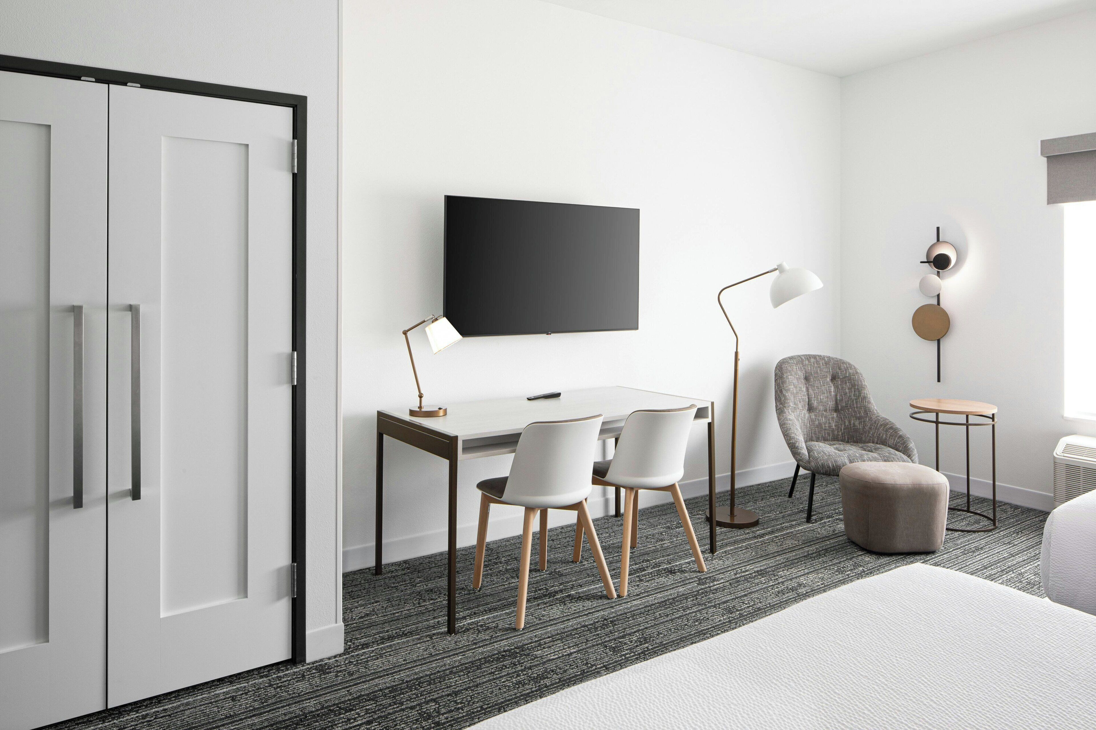 Habitación de hotel moderna con escritorio, dos sillas, un sillón gris, una mesita redonda y una cama.