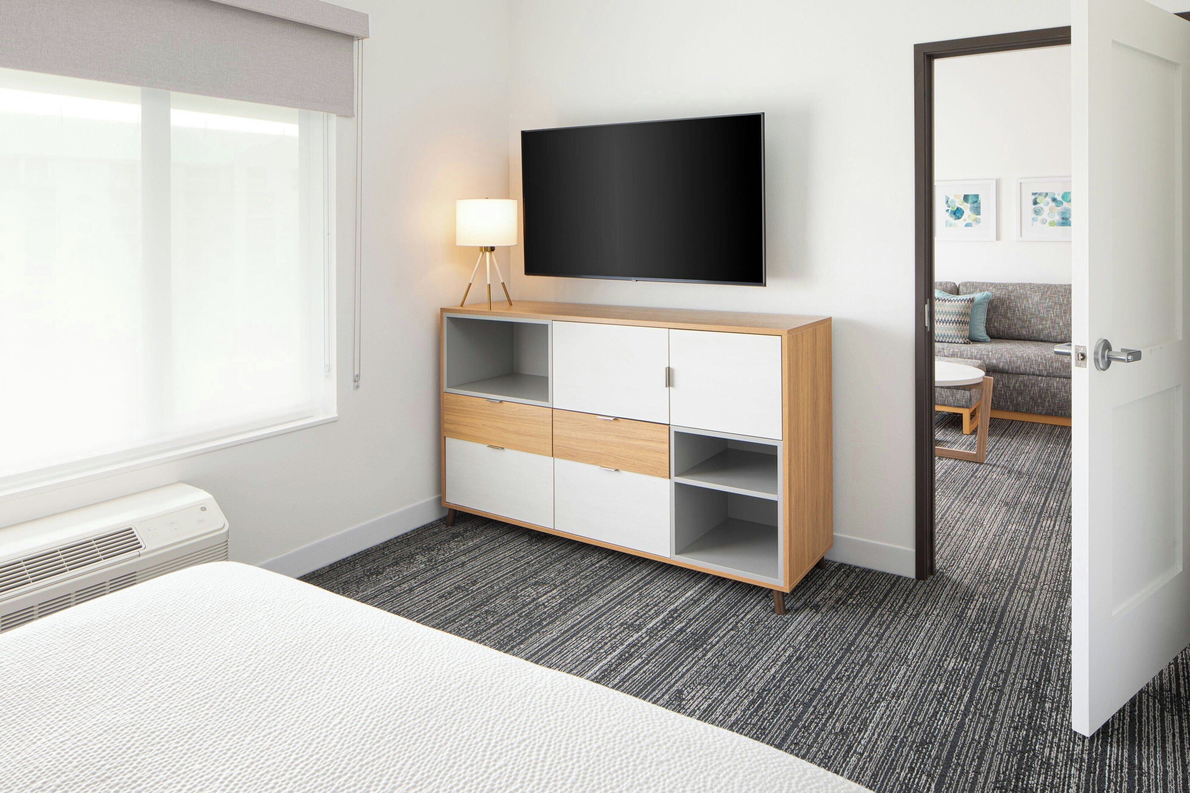 Una habitación de hotel con cama, televisor montado en la pared, mesita de noche con lámpara, armario y puerta que da a otra habitación.