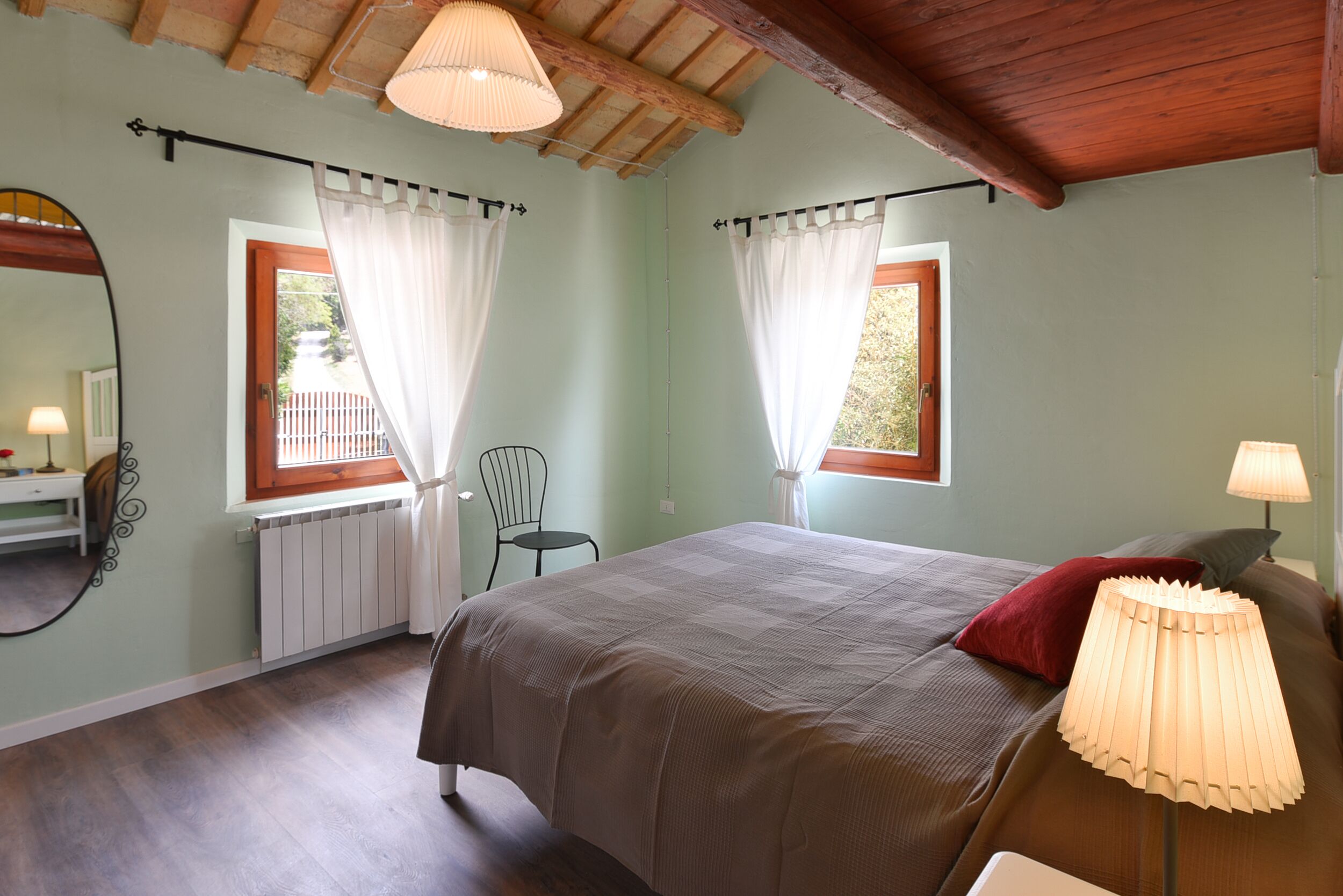 A bedroom with a bed, two windows with curtains, a chair, a lamp, and a radiator.