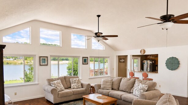 A spacious living room with a large window overlooking a lake, a ceiling fan, and a wooden coffee table. - Waterfront Cabin | Near ARK | Large Family Retreat (Warsaw)