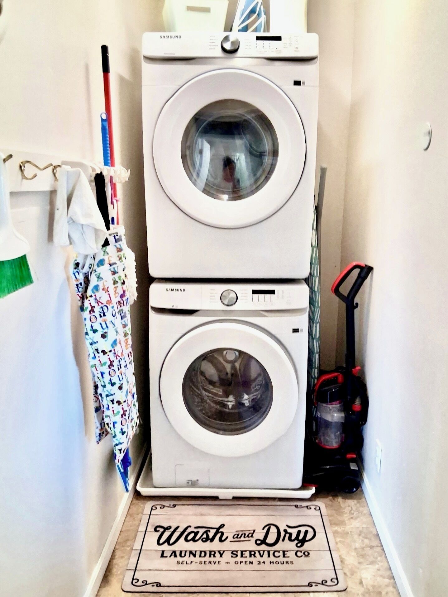 Pesuhuoneessa on kaksi päällekkäin asetettua pesukonetta, moppi, imuri ja pyykkiteline, jossa lukee: »Pese ja kuivaa».