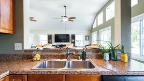A kitchen with granite countertops, a double sink, wooden cabinets, a ceiling fan, and a large window providing natural light. - Waterfront Cabin | Near ARK | Large Family Retreat (Warsaw)
