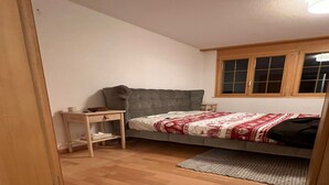 A small, single-bed room with a grey sofa, a wooden nightstand, and a window with wooden shutters. - Contemporary Swiss Escape (Gsteig)