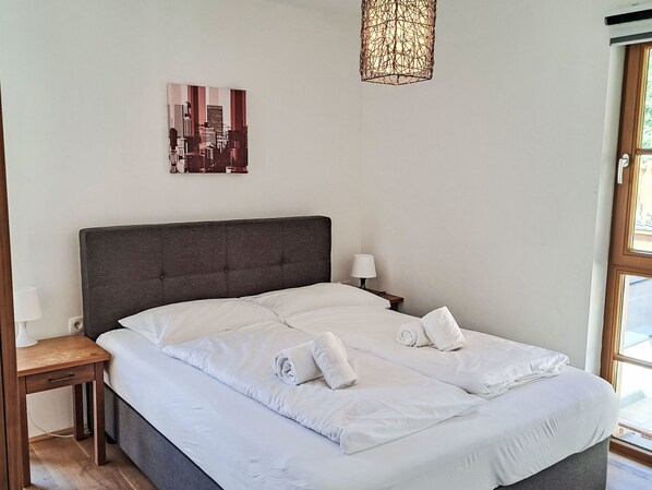 A neatly made bed with white linens and rolled towels, a dark headboard, a bedside lamp, and a cityscape artwork on the wall.