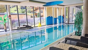 An indoor swimming pool with clear glass walls, wicker lounge chairs, and a view of a mountainous landscape.