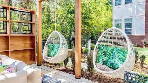 A patio with two hanging chairs, a wooden pergola, and a white building in the background. - Ellison on Broad (Greenville)