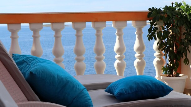 Un balcon doté d’une balustrade blanche, d’un pot de fleurs et d’un fauteuil avec des coussins bleus.