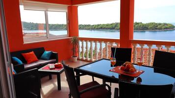 Un balcón con vista a un lago, una mesa con fruta y bebidas, y un sofá con cojines.