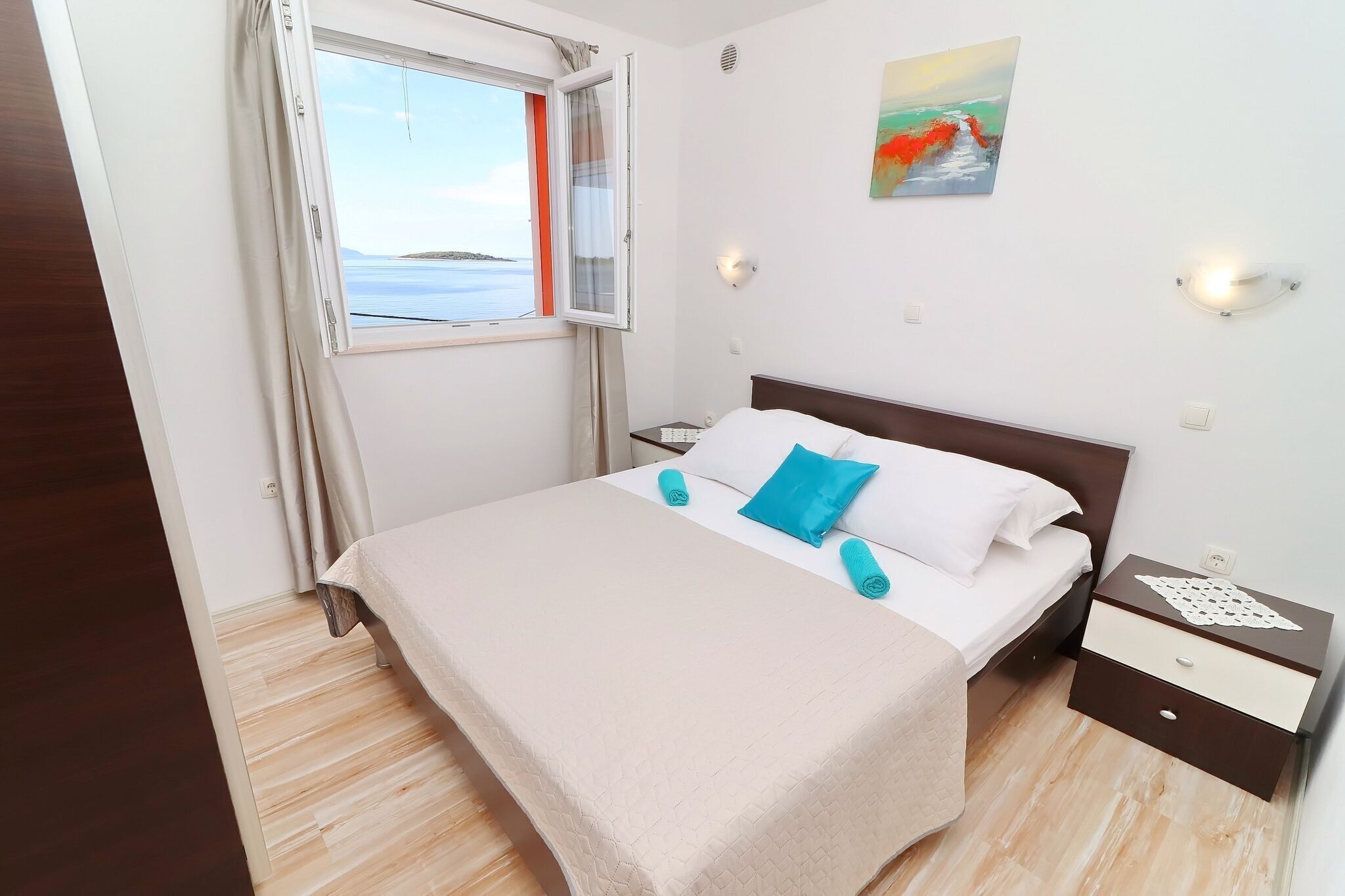 A neatly made bed with a beige bedspread and blue pillows, a small bedside table with a lamp, and a window with curtains.