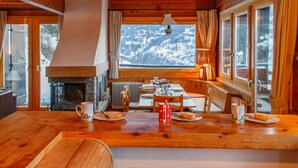 A cozy cabin interior with a wooden dining table, chairs, a fireplace, and large windows offering a view of snow-covered mountains. - Crettamour by Interhome (Veysonnaz)