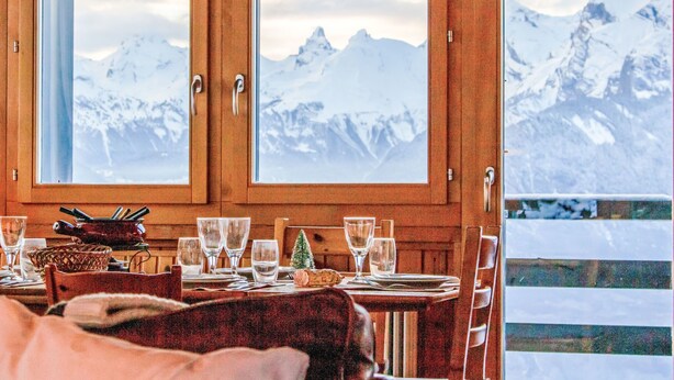 A dining area with a table set for two, wooden cabinets, and a view of snow-capped mountains through large windows. - Crettamour by Interhome (Veysonnaz)