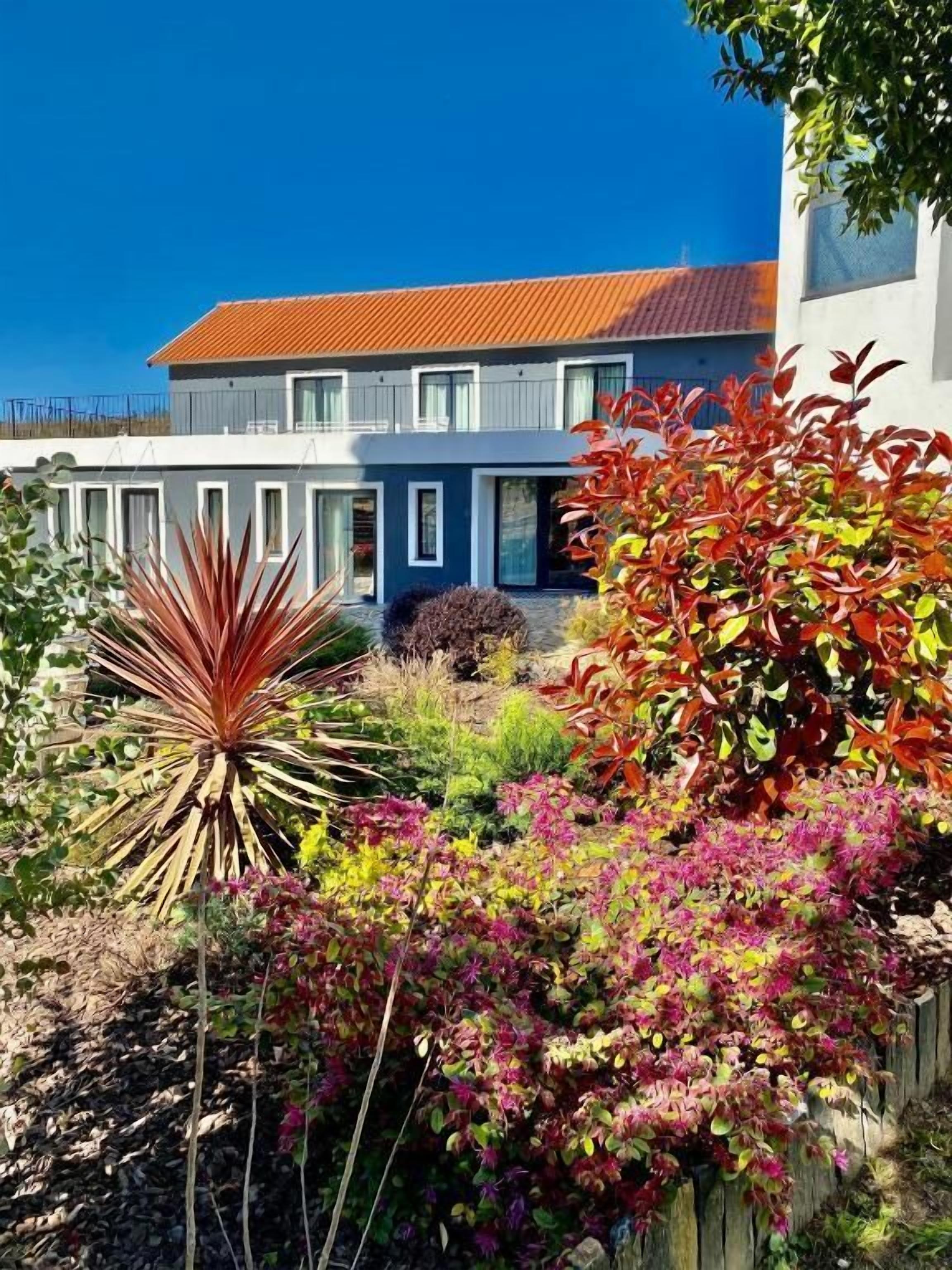 Um edifício moderno de dois andares com telhado de telhas vermelhas e um jardim bem cuidado com diversas plantas e arbustos.
