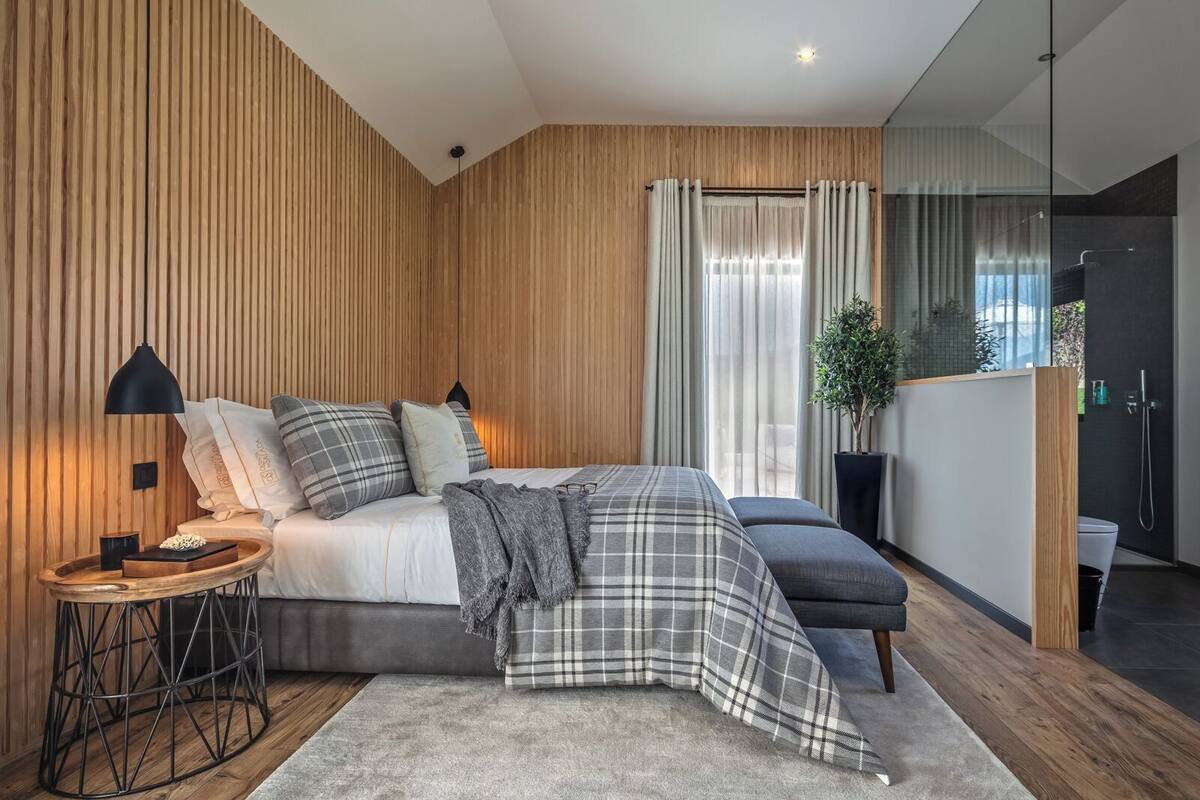 A modern bedroom with a wooden panel wall, a bed with plaid bedding, a side table with a lamp, and a view of a bathroom with a shower.