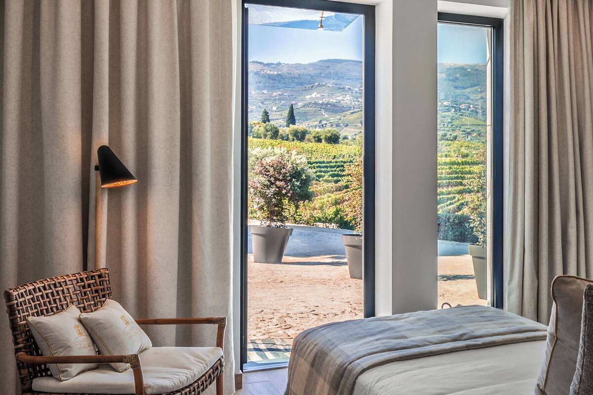 A bedroom with a large bed, a wicker chair, and a view of a vineyard through a glass door.