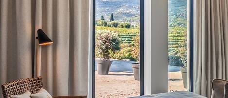 A bedroom with a large bed, a wicker chair, and a view of a vineyard through a glass door.