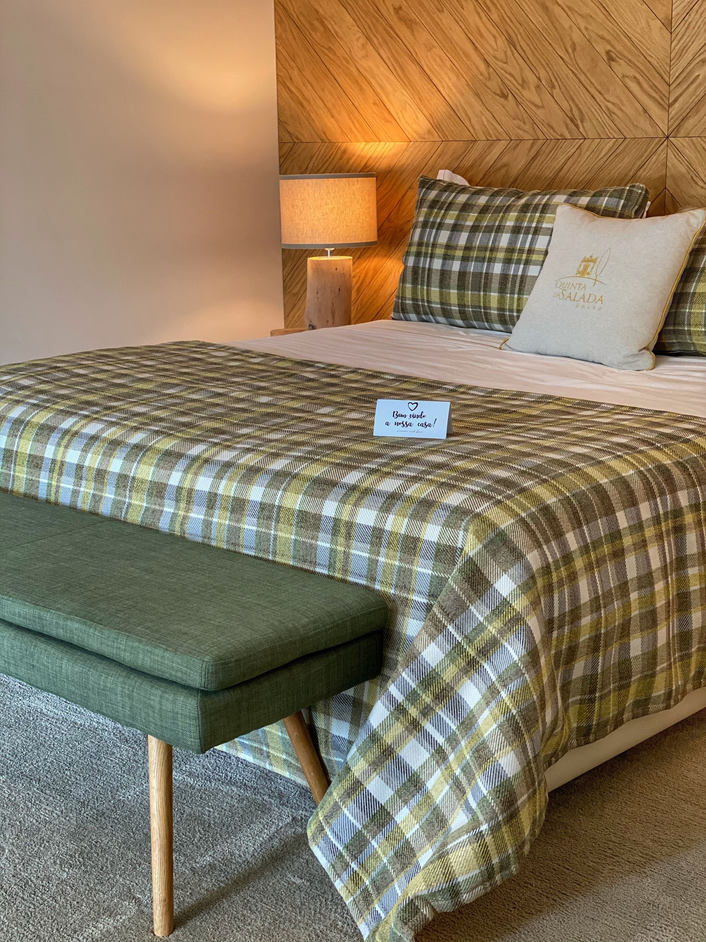 A neatly made bed with a checkered bedspread, a pillow with a logo, and a bedside lamp.