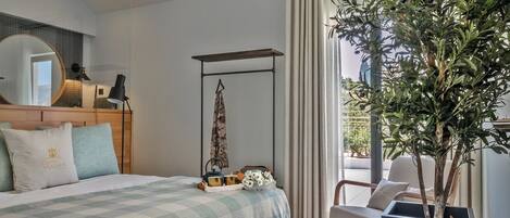 Un dormitorio con cama, mesita de noche, espejo, perchero, planta en maceta y vistas al exterior.