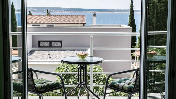 A balcony with two chairs and a table, overlooking a coastal view with buildings and trees. - Apartments Smokvica - One Bed/Apt with Balcony (Starigrad-Paklenica)