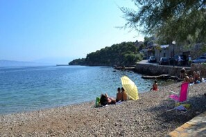 A pebbled beach with people relaxing under umbrellas, a stone wall, and a view of the sea. - Apartments St Rialto - Superior One Bedroom Apartment with Balcony (Jelsa)