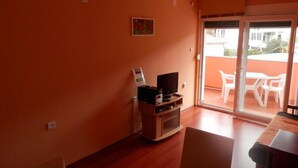 Une pièce comprenant un meuble de rangement en bois pour les appareils multimédias, un téléviseur et une porte-fenêtre coulissante donnant sur une terrasse extérieure avec des chaises blanches.