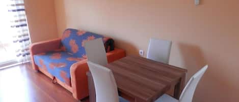A room with a wooden dining table, two white chairs, a sofa with a floral cover, and a window with blinds.